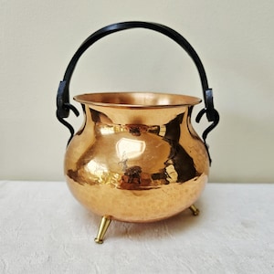 May include: A polished copper cauldron with a rounded body and a hammered texture. The vessel features a black arched metal handle attached to the sides and stands on three small, tapered brass-coloured legs. The rim is smooth and flared outward.