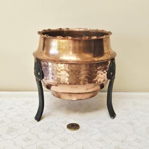 May include: A hammered copper bowl with a scalloped rim and three black, curved legs. The bowl has a dark patina and is likely vintage. A coin is on the surface in front of the bowl.