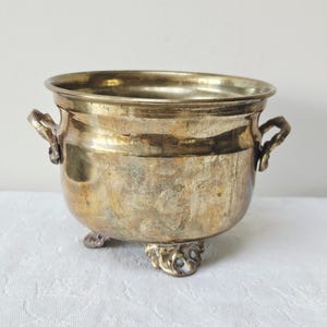 Può includere: Un vaso rotondo in ottone con finitura lucida, dotato di due piccole maniglie laterali e tre piedi decorativi ornati. La superficie metallica presenta variazioni naturali di colore e consistenza, con un'ampia apertura superiore e un corpo curvo.