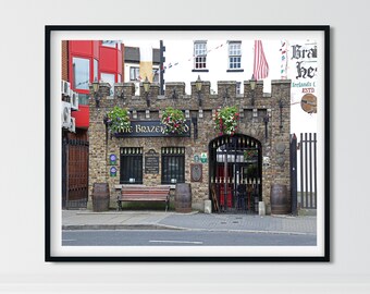 May include: A brick building with a black iron gate and a sign that reads "The Brazen Head" above the entrance. The building has a red brick facade and a green door. There are two barrels in front of the building.