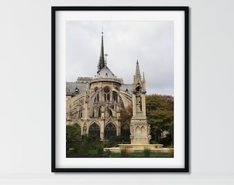 May include: A black and white photo of the Notre Dame Cathedral in Paris, France. The photo shows the back of the cathedral with a tall spire and intricate stonework. There is a small fountain in the foreground.