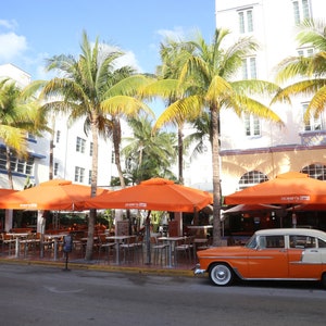 May include: A vintage orange and cream car parked in front of a restaurant with orange umbrellas and palm trees.
