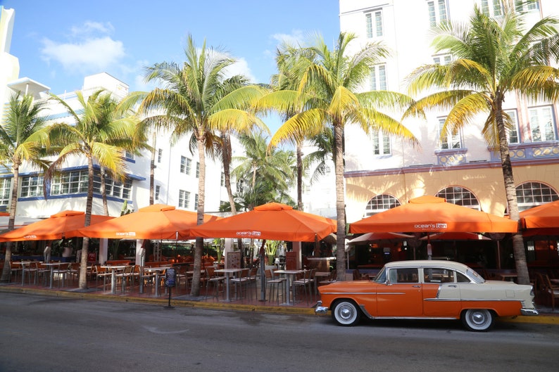 May include: A vintage orange and cream car parked in front of a restaurant with orange umbrellas and palm trees.