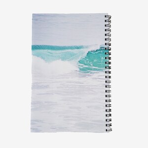 May include: Spiral-bound notebook featuring a serene ocean wave design. The cover showcases a turquoise wave cresting over white water, set against a pale blue sky. The notebook's binding is black.