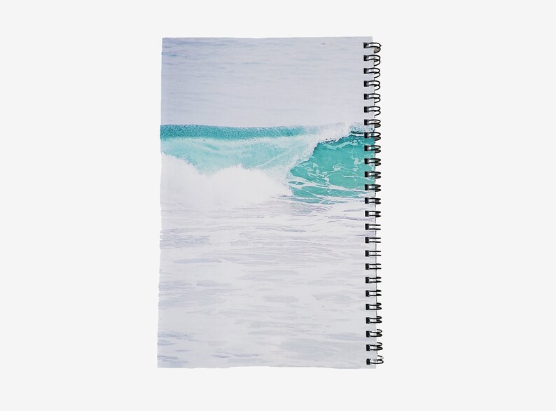 May include: Spiral-bound notebook featuring a serene ocean wave design. The cover showcases a turquoise wave cresting over white water, set against a pale blue sky. The notebook's binding is black.