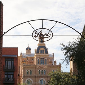 May include: A black metal archway with the word "Pearl" in a white oval sign. The archway is in front of a brick building with a sign that reads "San Antonio Brewery No. 1".