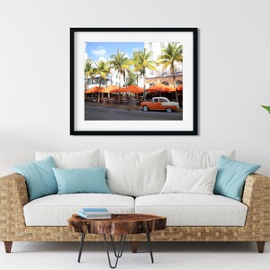 May include: A vintage orange and white car parked in front of a restaurant with orange umbrellas and palm trees. The restaurant is located on a street with a white building in the background.