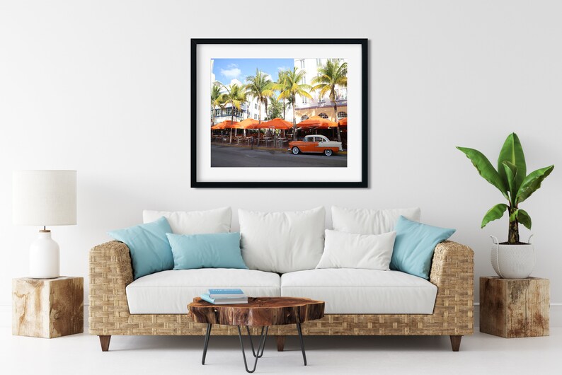 May include: A vintage orange and white car parked in front of a restaurant with orange umbrellas and palm trees. The restaurant is located on a street with a white building in the background.
