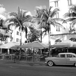 May include: A black and white photograph of a vintage car parked in front of a building with palm trees. The car is a classic American saloon with a chrome bumper and whitewall tyres. The building is a two-storey structure with a tiled roof and large windows. The palm trees are tall and slender, with lush green fronds.