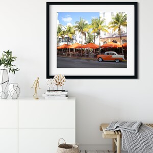 May include: A vintage car parked in front of a building with orange umbrellas and palm trees. The car is white with orange accents and has a black roof. The building is white with blue accents and has a sign that says "The Living Room".