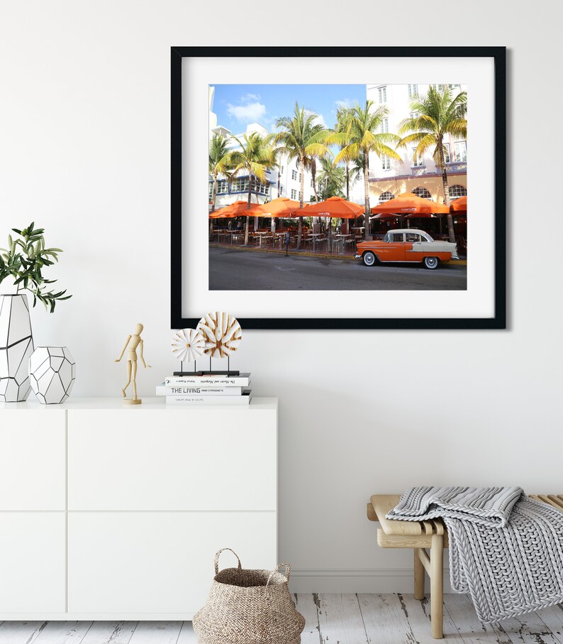May include: A vintage car parked in front of a building with orange umbrellas and palm trees. The car is white with orange accents and has a black roof. The building is white with blue accents and has a sign that says "The Living Room".