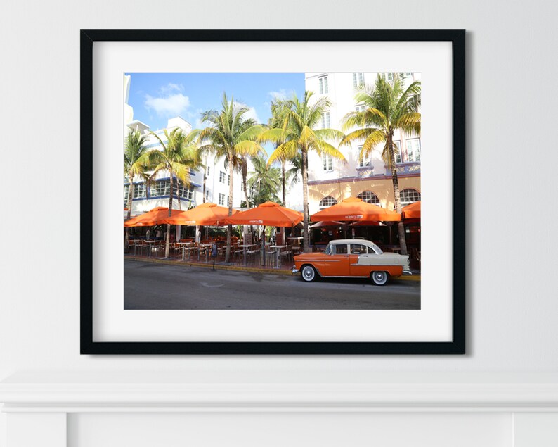 May include: A vintage orange and white car parked in front of a restaurant with orange umbrellas and palm trees.
