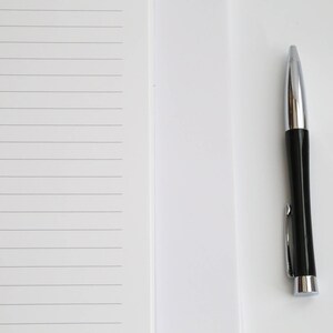 May include: A black and silver pen rests on a white surface next to a lined notepad. The pen has a shiny silver tip and a black barrel with a silver clip. The notepad has horizontal gray lines.