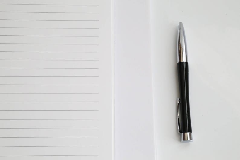 May include: A black and silver pen rests on a white surface next to a lined notepad. The pen has a shiny silver tip and a black barrel with a silver clip. The notepad has horizontal gray lines.