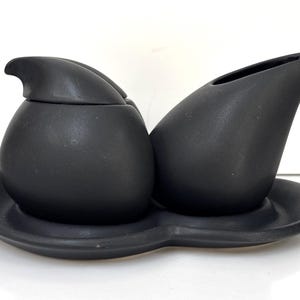 May include: A matte black ceramic creamer and sugar bowl set with a matching oval tray. The sugar bowl features a rounded lid with a small handle, while the creamer has a curved pouring spout. Both pieces sit securely on the contoured base.