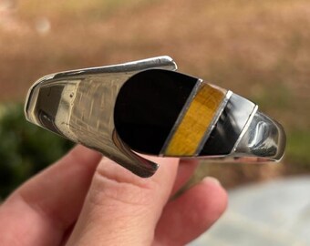 May include: A silver-toned cuff bracelet with a geometric design. The bracelet features a large black oval, a yellow stripe, and a silver accent. The bracelet is held in a hand.