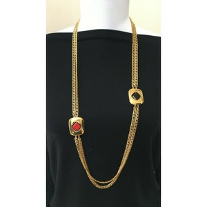 May include: A long gold-tone necklace with a chain design. The necklace features two square pendants, one with a red center and the other with a black center. The necklace is displayed on a black background.