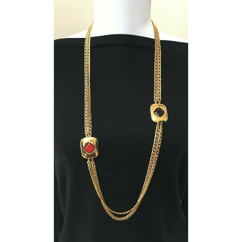 May include: A long gold-tone necklace with a chain design. The necklace features two square pendants, one with a red center and the other with a black center. The necklace is displayed on a black background.