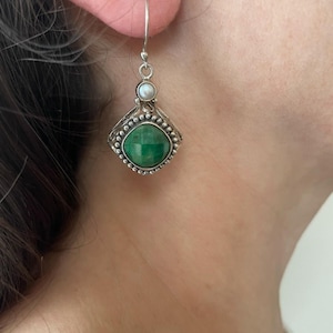 May include: A silver earring with a green gemstone and a small white pearl. The gemstone is square-shaped with a faceted surface, set in a decorative silver frame. The earring hangs from a silver hook.