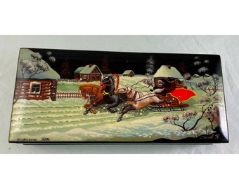 May include: A black lacquered wooden box with a painted scene of a horse-drawn sleigh in a snowy winter village. The box is decorated with a red, white, and blue flag and the text "Фрязино 1955".