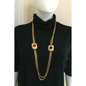 May include: Gold-tone chain necklace with two square pendants. One pendant features a red center, the other a black center. The necklace is displayed on a black turtleneck.