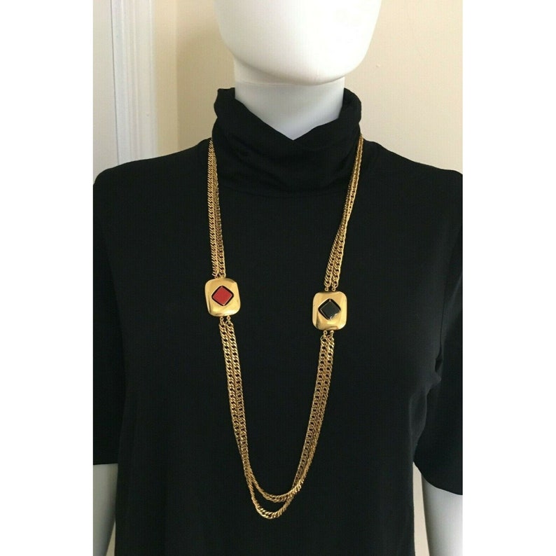 May include: Gold-tone chain necklace with two square pendants. One pendant features a red center, the other a black center. The necklace is displayed on a black turtleneck.