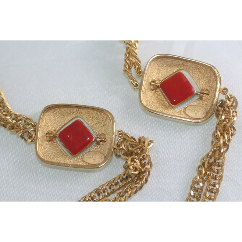 May include: Gold-tone necklace featuring two square pendants with a red diamond-shaped center. The pendants are connected by a gold chain. The necklace has a vintage aesthetic and the brand name "VALENTINO" is visible.