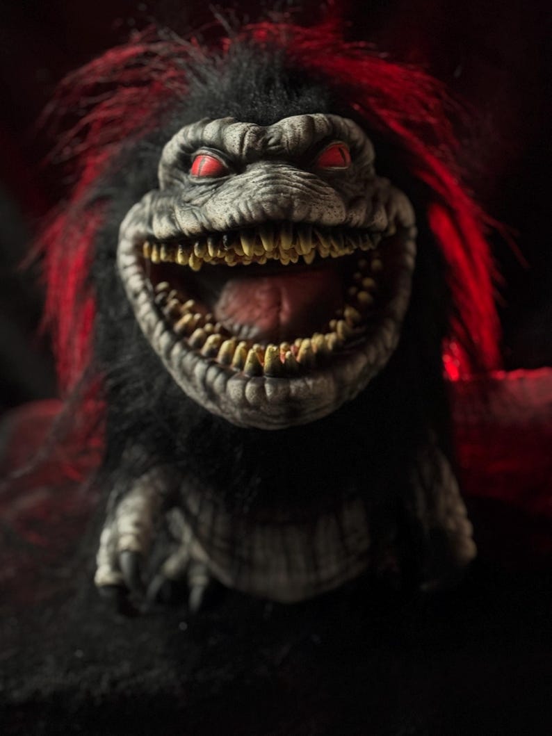 May include: A close-up of a creature with a menacing grin, featuring sharp teeth and red eyes. The creature has black fur with red streaks, and a textured, gray face. The image is dark, creating a spooky atmosphere.