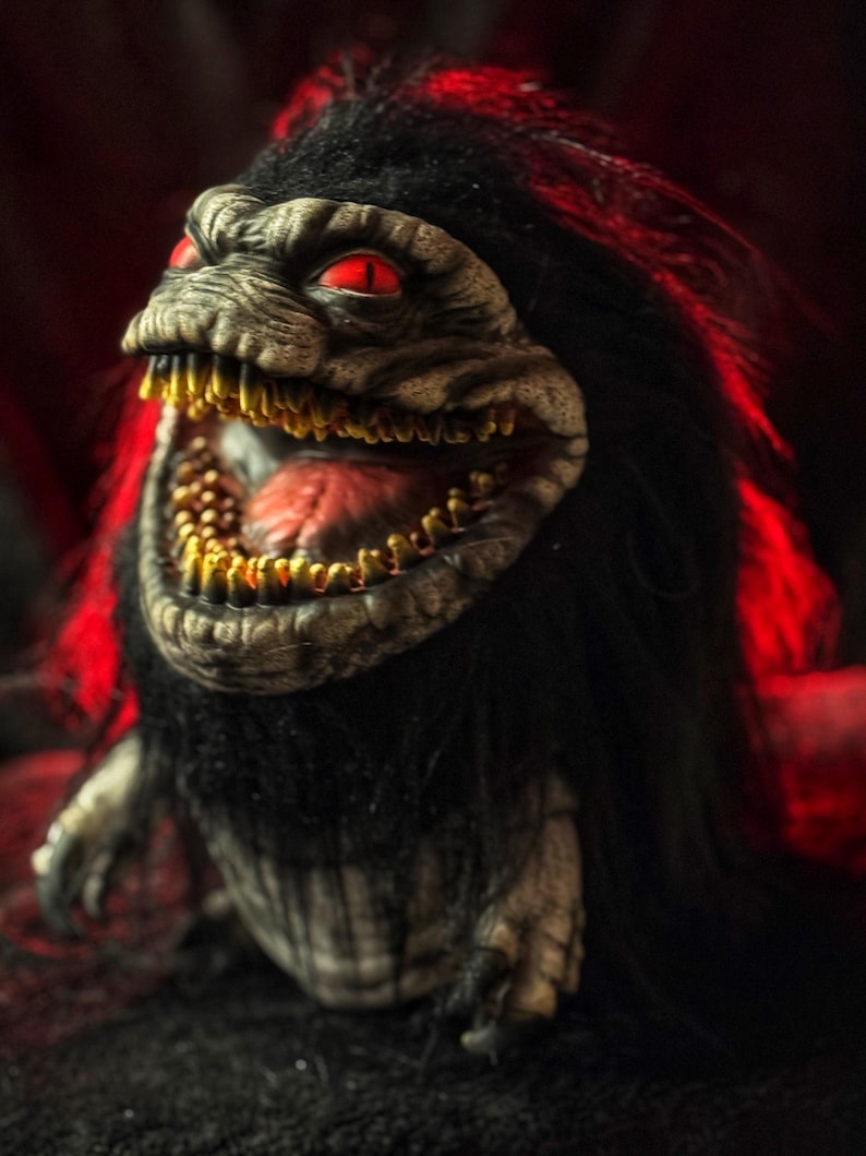 May include: A detailed close-up of a Critters creature, featuring a textured, gray face with red eyes and a wide-open mouth revealing rows of golden teeth. The creature has black fur and is set against a dark, red-tinged background.