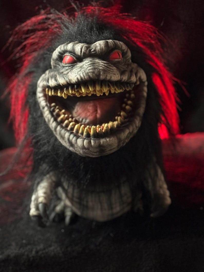 May include: A close-up of a creature with a wide, toothy grin and red eyes. The creature has black fur and red highlights. Its skin is gray and textured, with a large mouth filled with yellow teeth. The overall appearance is menacing.