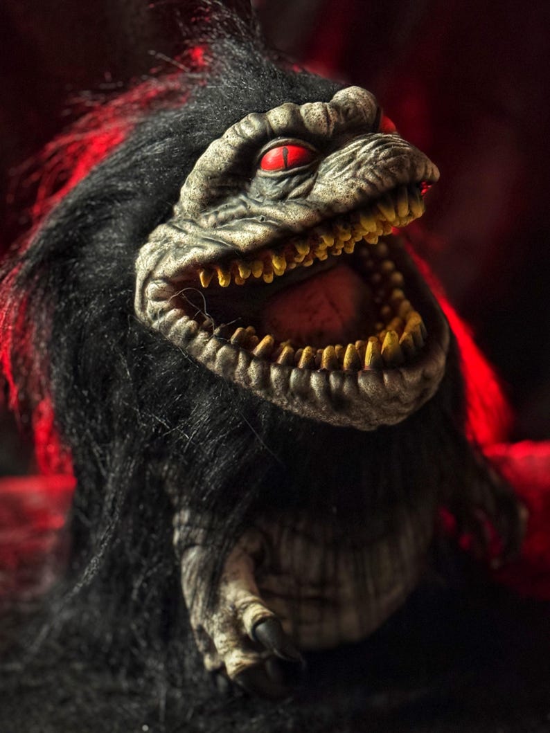 May include: A close-up of a creature with a reptilian face, red eyes, and a wide-open mouth revealing yellow teeth. The creature has black fur and a textured, gray face. The background is a dark red.