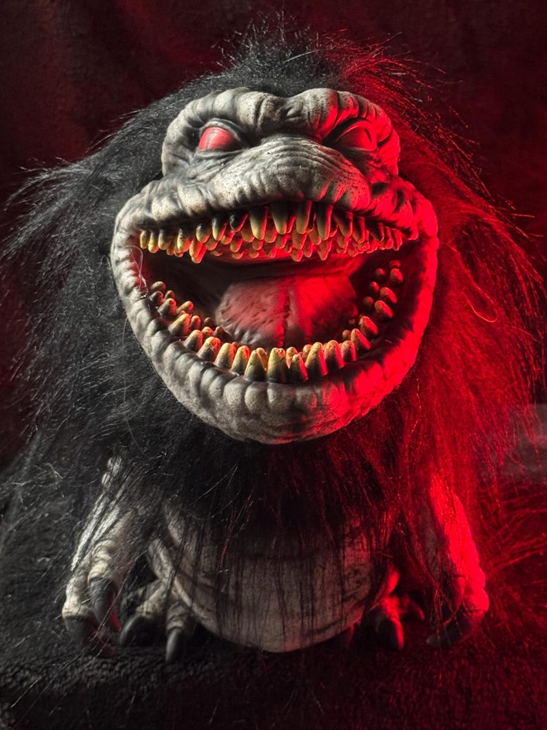 May include: A close-up of a monster with a gray face, red eyes, and a wide, toothy grin. The monster has black fur and is illuminated with red light, creating a spooky and menacing effect. The creature's teeth are yellow and sharp.