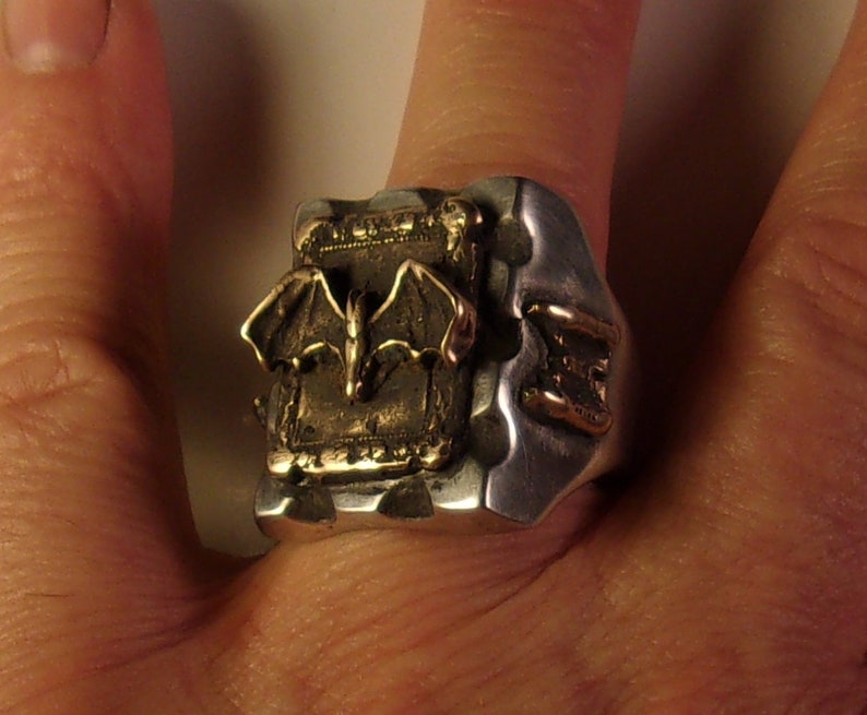 Puede incluir: Anillo de plata con un dise&ntilde;o detallado de murci&eacute;lago en color bronce. El anillo tiene una apariencia texturizada y una est&eacute;tica g&oacute;tica &uacute;nica. El murci&eacute;lago est&aacute; centrado, con las alas extendidas. Una pieza de joyer&iacute;a llamativa.