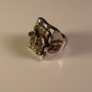 Puede incluir: Anillo plateado con superficie texturizada y detalles dorados. El anillo presenta un emblema dorado con un dise&ntilde;o de murci&eacute;lago y otros adornos dorados. Una pieza de joyer&iacute;a &uacute;nica.