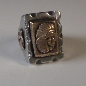Puede incluir: Un anillo de plata con una superficie texturizada y una incrustación rectangular dorada que representa a un jefe nativo americano con un tocado.