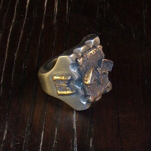 Puede incluir: Un anillo plateado con un dise&ntilde;o texturizado y detalles dorados. El anillo presenta las letras "E" y "A" en oro. El anillo tiene una apariencia robusta y art&iacute;stica.