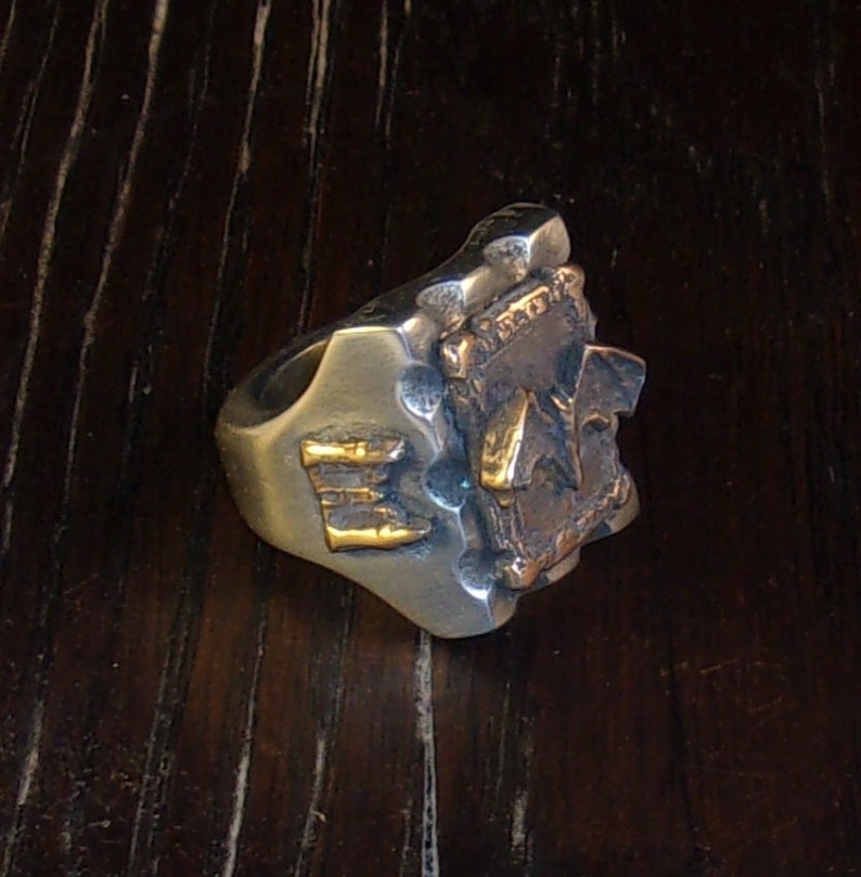 Puede incluir: Un anillo plateado con un dise&ntilde;o texturizado y detalles dorados. El anillo presenta las letras "E" y "A" en oro. El anillo tiene una apariencia robusta y art&iacute;stica.