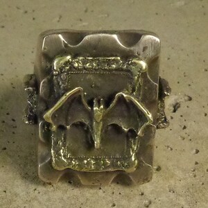 Puede incluir: Anillo plateado con una cara rectangular. La cara presenta un dise&ntilde;o de murci&eacute;lago dorado dentro de un marco decorativo. El anillo tiene un aspecto texturizado y ligeramente desgastado.