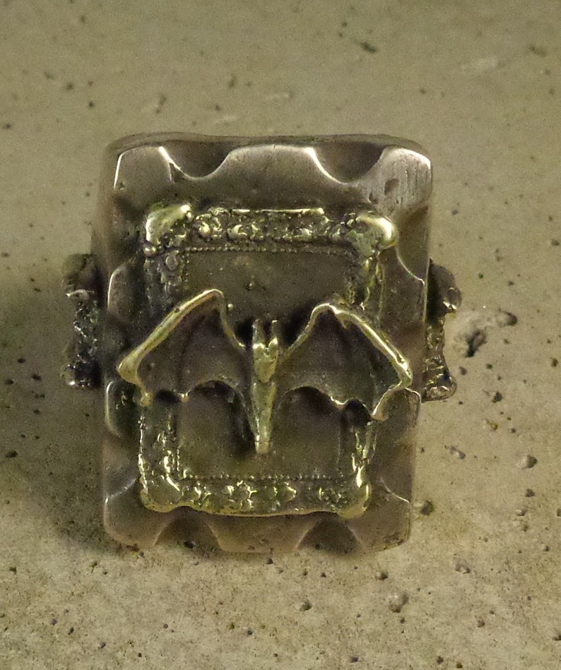 Puede incluir: Anillo plateado con una cara rectangular. La cara presenta un dise&ntilde;o de murci&eacute;lago dorado dentro de un marco decorativo. El anillo tiene un aspecto texturizado y ligeramente desgastado.