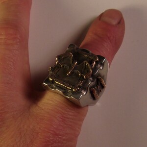 Puede incluir: Un anillo plateado con un dise&ntilde;o geom&eacute;trico texturizado y detalles dorados. El anillo se lleva en un pulgar, mostrando su forma abstracta &uacute;nica y elementos met&aacute;licos contrastantes.