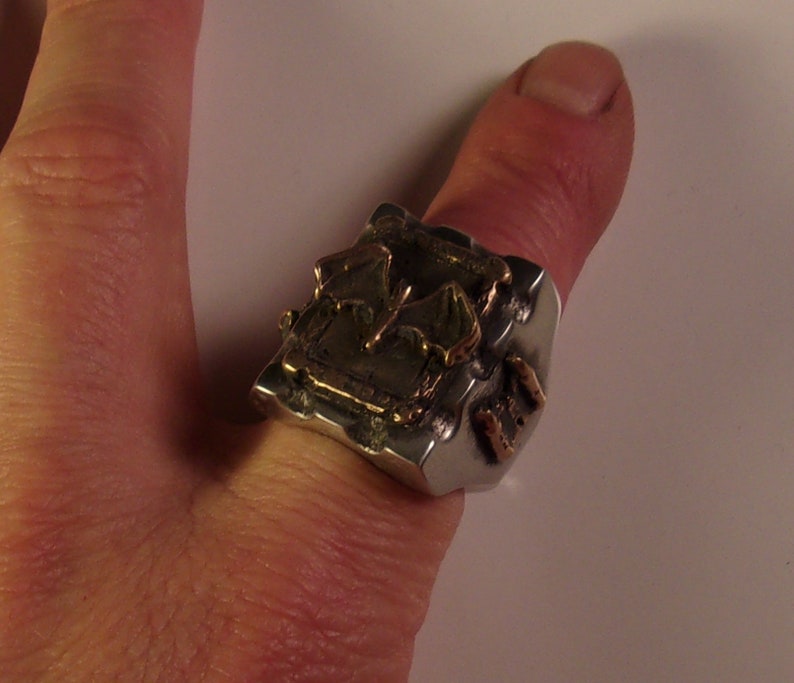 Puede incluir: Un anillo plateado con un dise&ntilde;o geom&eacute;trico texturizado y detalles dorados. El anillo se lleva en un pulgar, mostrando su forma abstracta &uacute;nica y elementos met&aacute;licos contrastantes.