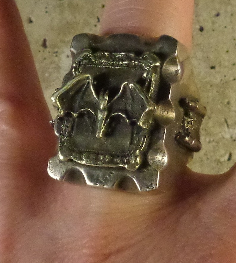 Puede incluir: Un anillo plateado con un dise&ntilde;o g&oacute;tico detallado. El anillo presenta un motivo central de dos alas de murci&eacute;lago estilizadas y elementos arquitect&oacute;nicos. El anillo se lleva en un dedo, mostrando su intrincada artesan&iacute;a y est&eacute;tica &uacute;nica.