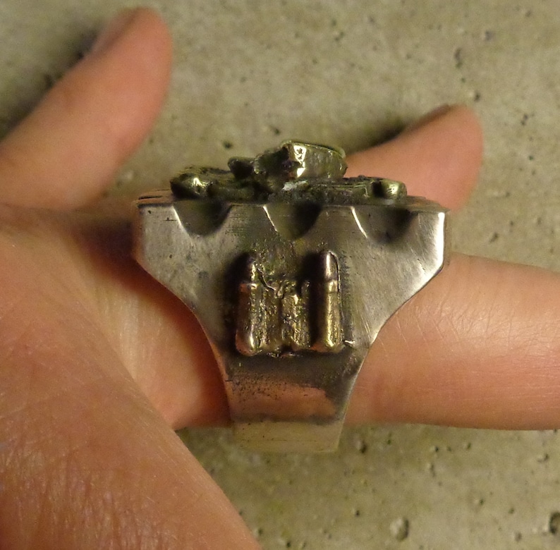 Puede incluir: Un anillo plateado con una cara rectangular. El anillo presenta un dise&ntilde;o en relieve y texturizado en tono dorado, posiblemente representando un castillo o un elemento arquitect&oacute;nico. El anillo est&aacute; sostenido por una mano.