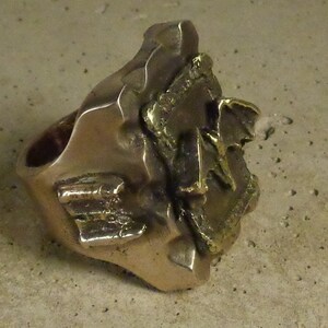 Puede incluir: Un anillo de color bronce con un dise&ntilde;o abstracto y texturizado. El anillo presenta una banda gruesa y una parte superior grande, de forma irregular, con detalles en relieve. La superficie tiene un aspecto ligeramente desgastado.