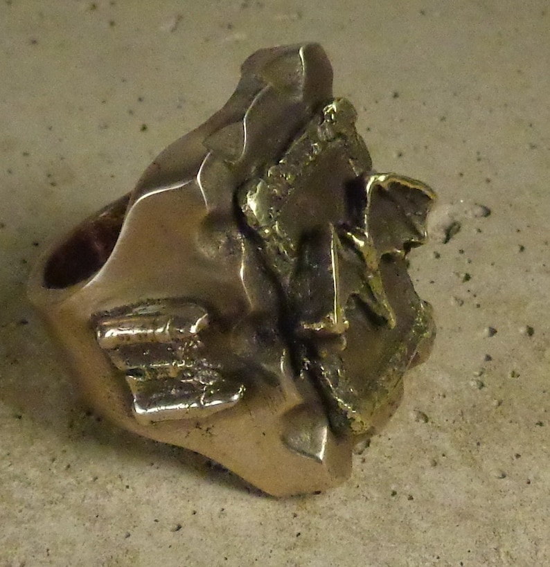 Puede incluir: Un anillo de color bronce con un dise&ntilde;o abstracto y texturizado. El anillo presenta una banda gruesa y una parte superior grande, de forma irregular, con detalles en relieve. La superficie tiene un aspecto ligeramente desgastado.