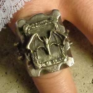 Puede incluir: Anillo plateado con dise&ntilde;o de murci&eacute;lago. El anillo tiene una cara rectangular con un dise&ntilde;o de marco en relieve y un murci&eacute;lago en el centro. El anillo se lleva en un dedo.