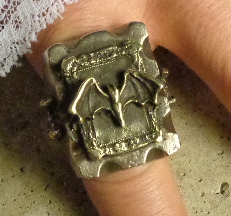Puede incluir: Anillo plateado con dise&ntilde;o de murci&eacute;lago. El anillo tiene una cara rectangular con un dise&ntilde;o de marco en relieve y un murci&eacute;lago en el centro. El anillo se lleva en un dedo.