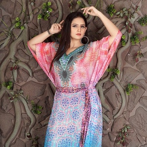 May include: A long, flowing pink and blue patterned kaftan with a V-neck and a belt. The kaftan has a colourful geometric design on the bodice and a floral pattern on the skirt.