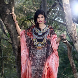 Puede incluir: Una mujer con una kaftán de estampado rosa y negro con un collar de cuentas. La kaftán tiene un escote en V y es fluida. La mujer está de pie en un entorno de bosque.