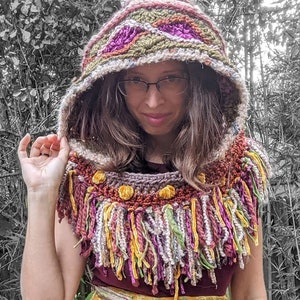 May include: A woman wearing a colourful crocheted hooded scarf with fringe and a yellow floral wrap skirt.